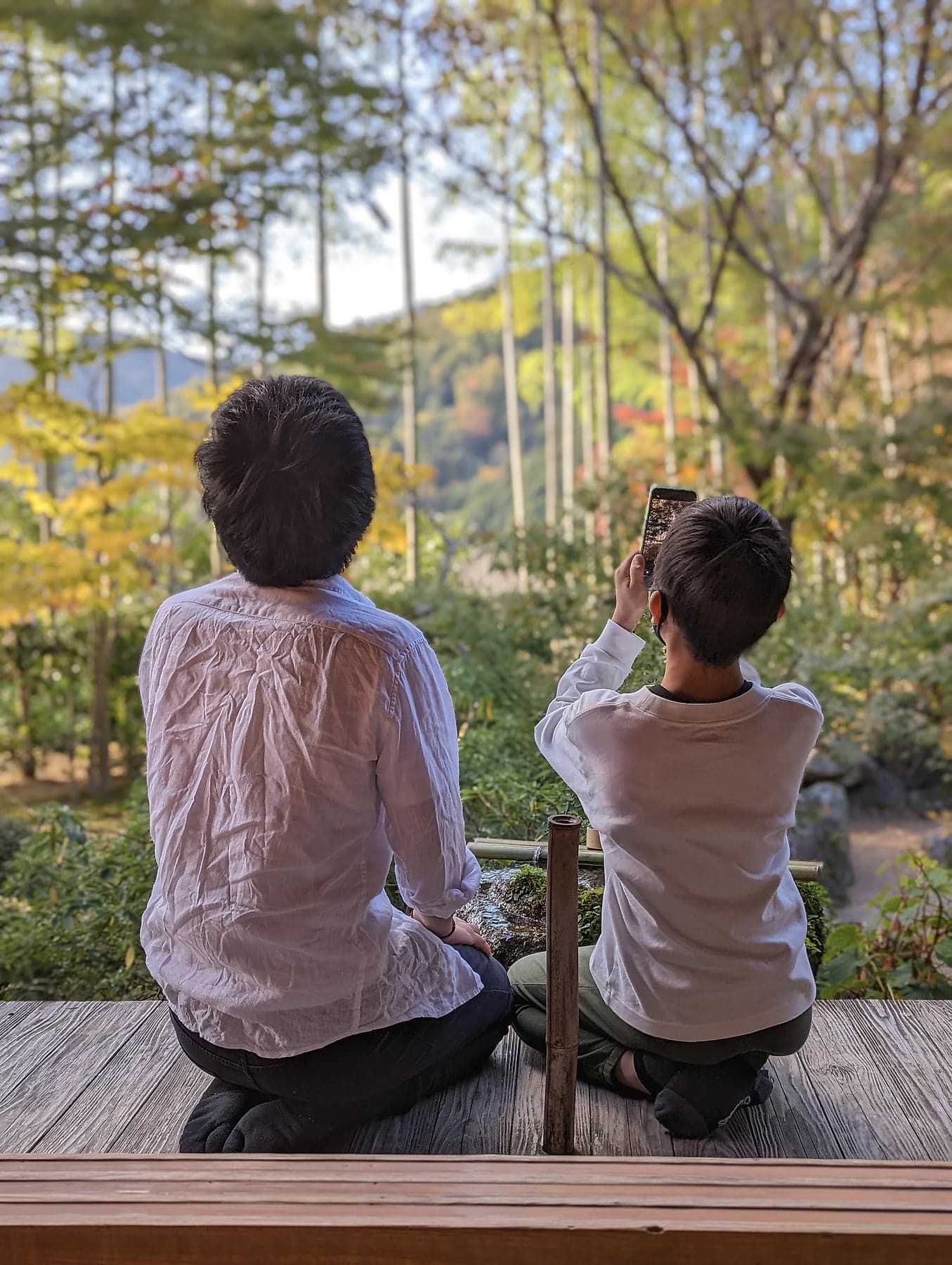 A father and son sitting on a wooden engawa looking out at bamboo and autumn foliage in a temple garden