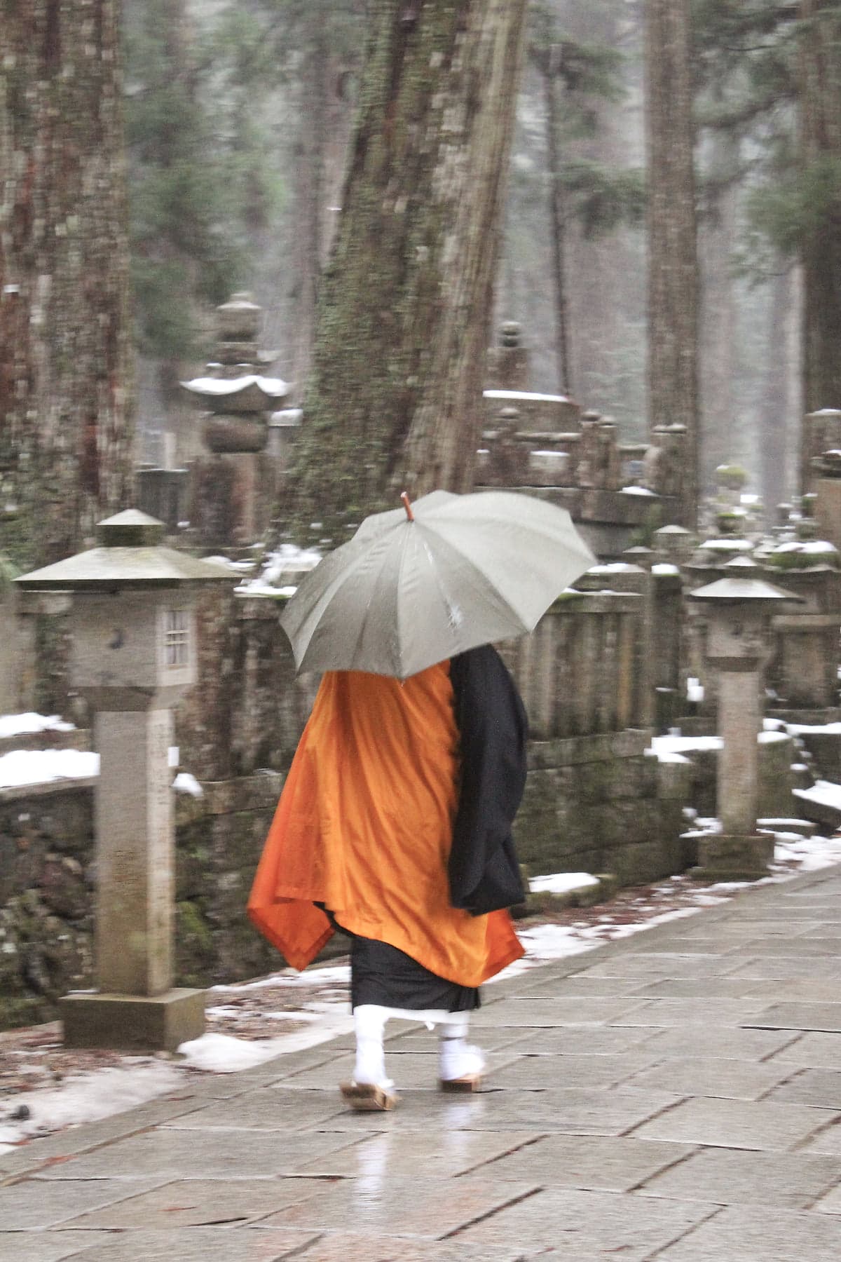 A monk in orange kesa and black robes walking with an umbrella through Okunoin cemetery in light rain, stone lanterns and cedars on either side