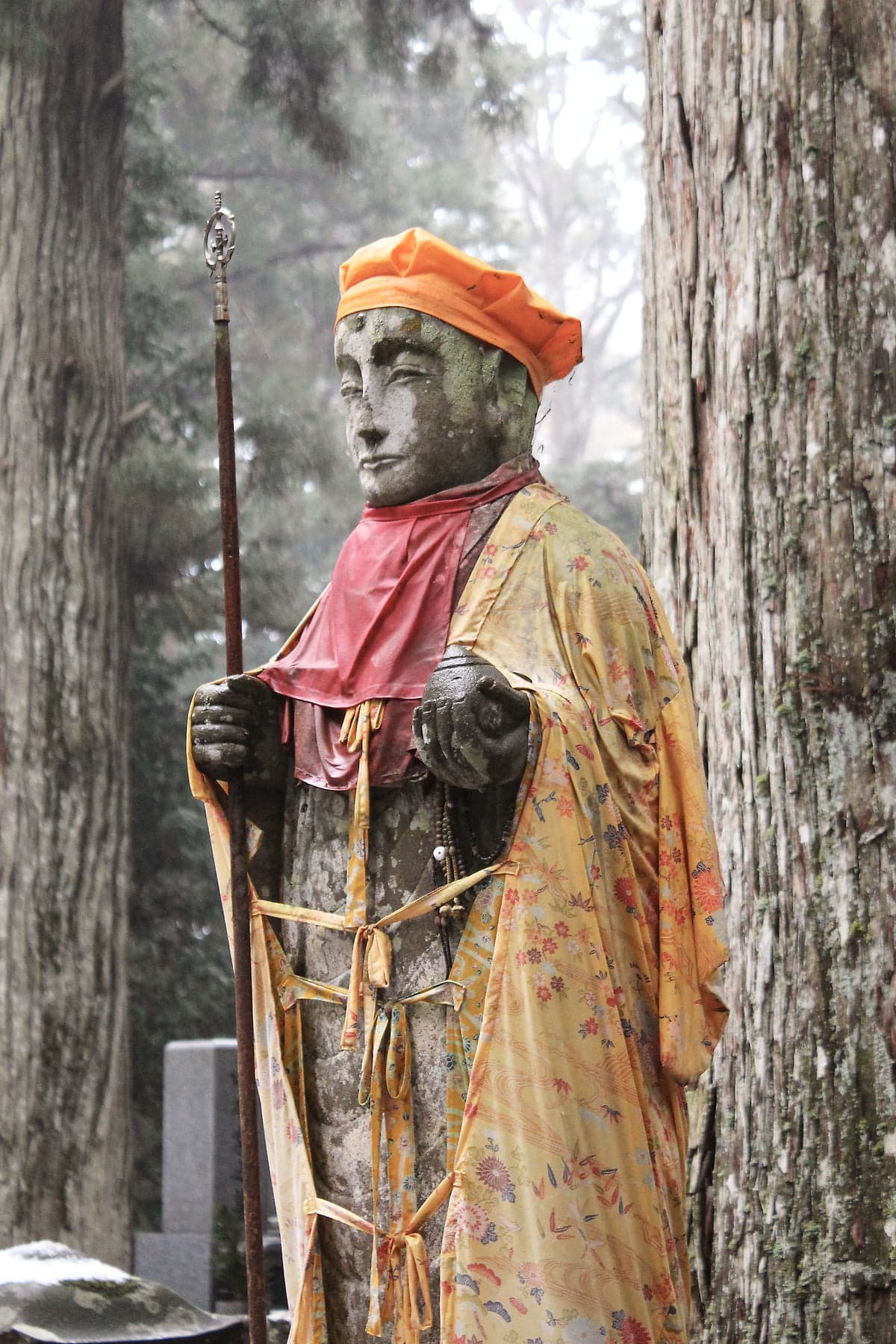 A stone statue dressed in an orange cap, pink bib, and floral kimono, holding a staff among the cedars and graves of Okunoin