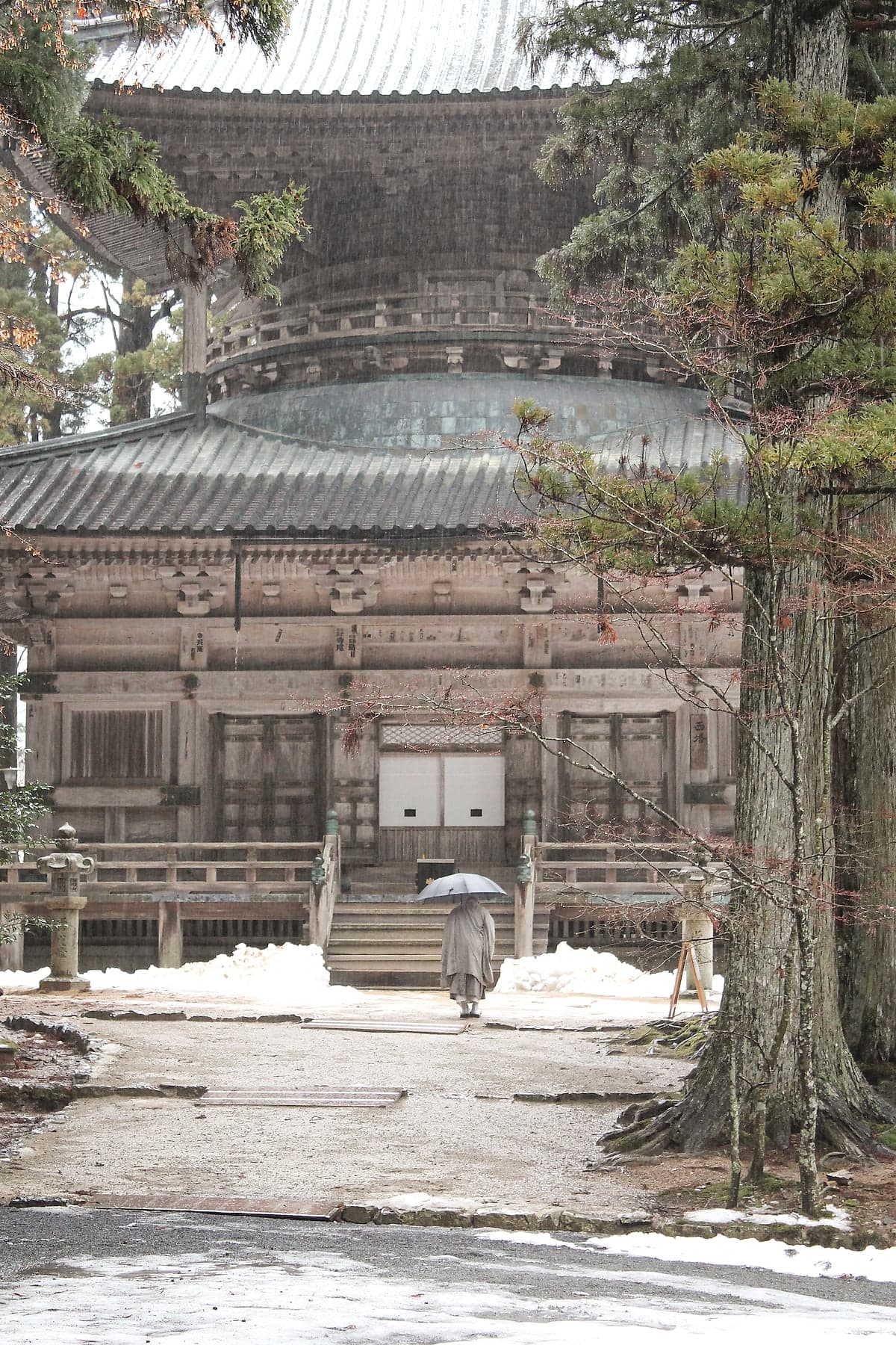 A figure with an umbrella approaching a weathered wooden temple building on Koya-san, snow banked on either side of the path, ancient cedars framing the scene