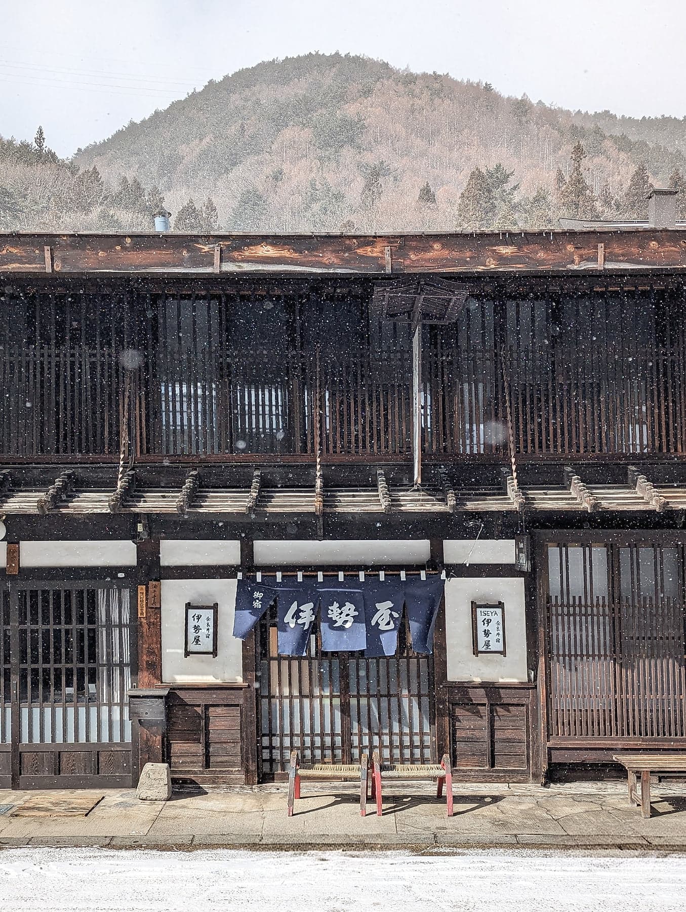 A latticed wooden facade with indigo noren curtains and snow falling against dark timber in Narai-juku