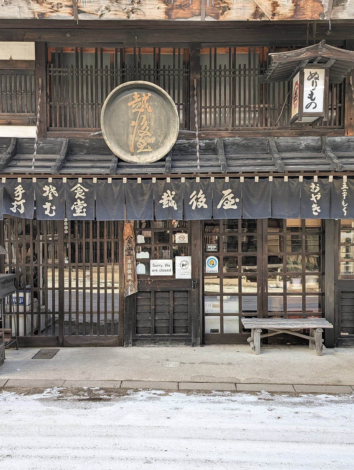 A traditional shopfront with a round wooden sign, dark noren curtains, and a weathered bench in Narai-juku