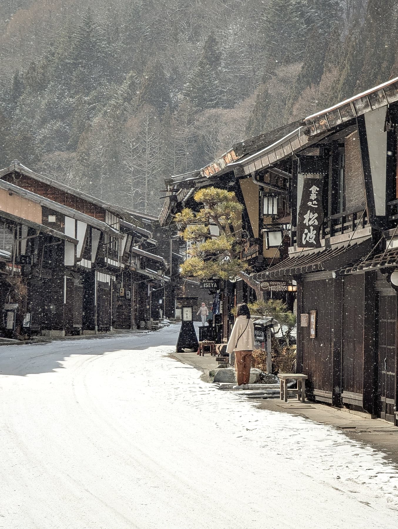 A lone figure walking through heavy snowfall past dark wooden buildings and a pine tree in Narai-juku
