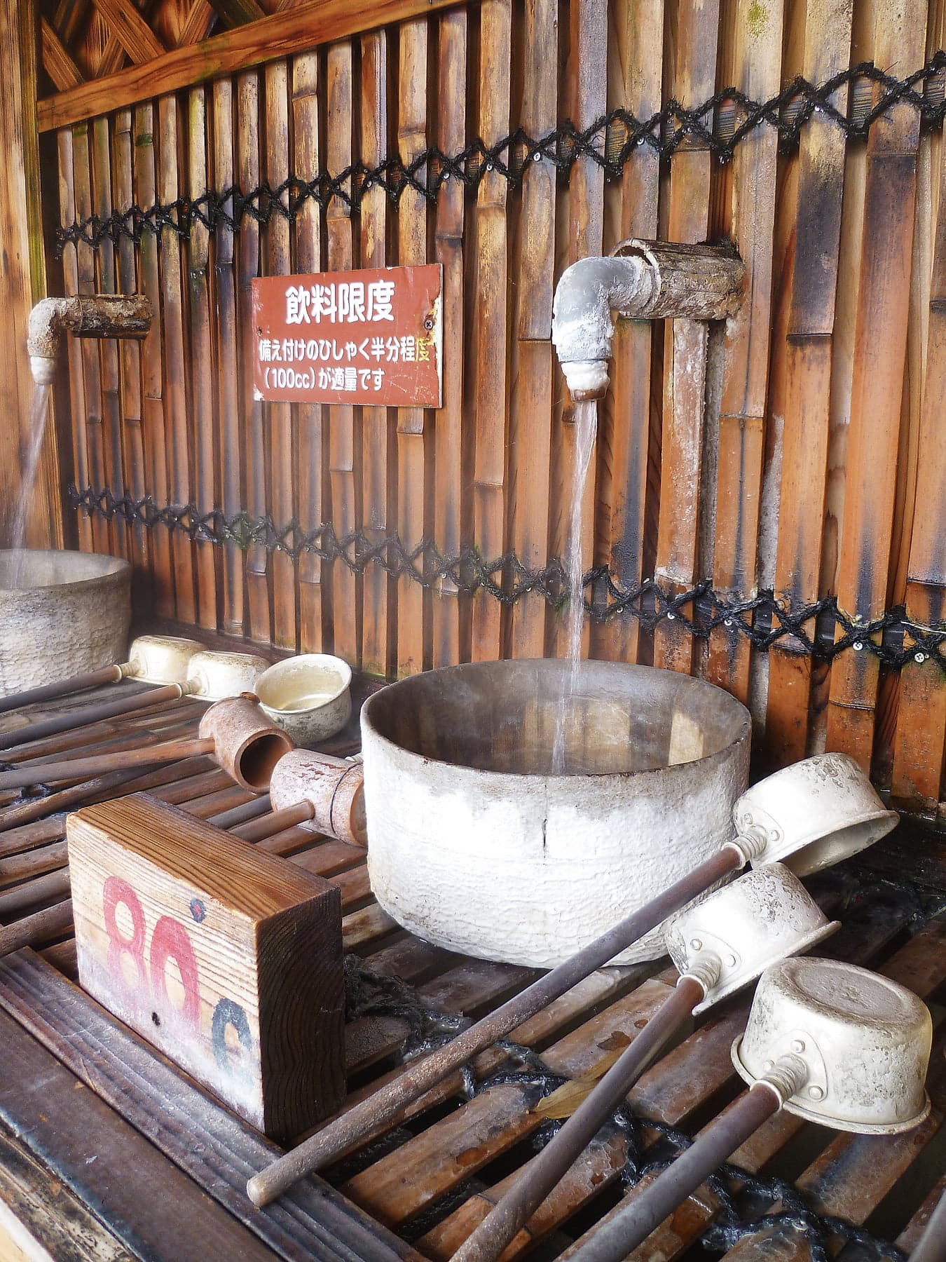 Wooden ladles and a metal basin at an onsen drinking water station, steam rising, bamboo wall behind