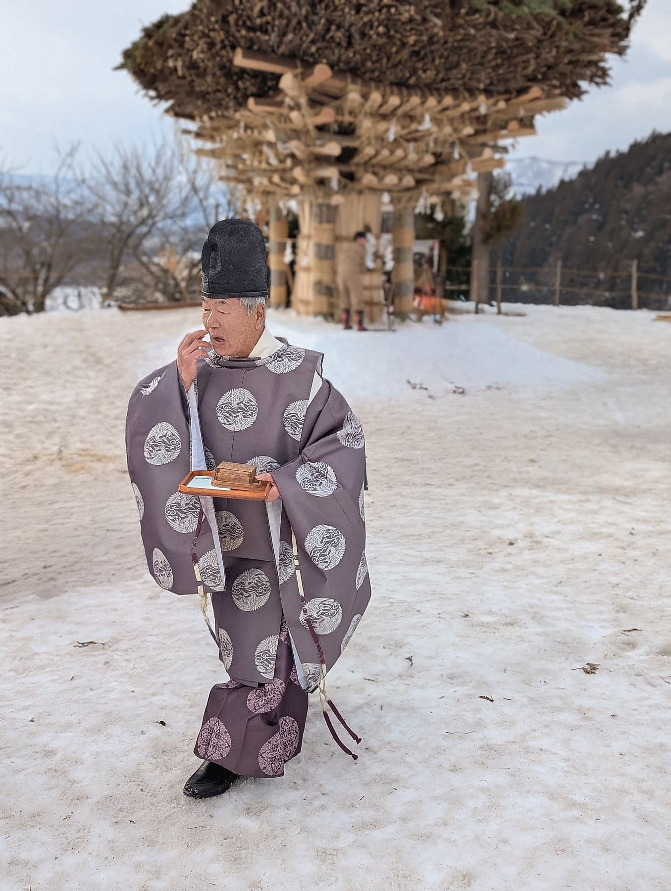 A Shinto priest in ceremonial robes walking across the snow at Nozawa Onsen, the wooden shaden shrine rising behind him