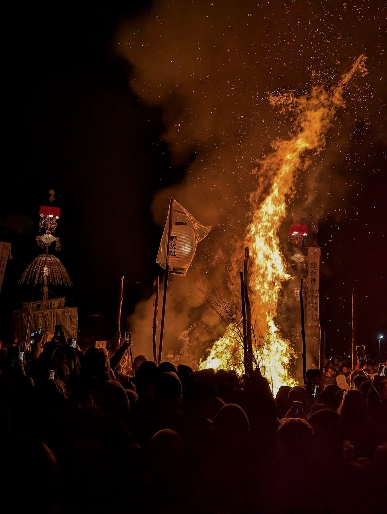 A tower of flame and sparks rising into the night sky above the crowd at Nozawa Onsen's Dosojin Matsuri, the dosojin pillar silhouetted to the left