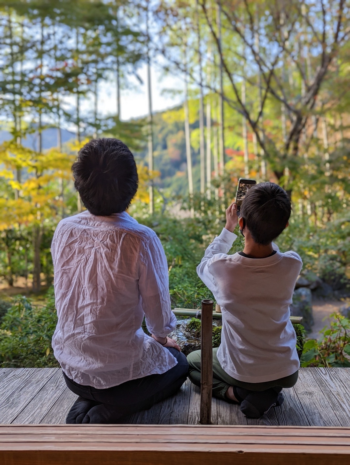 Two people sitting on a wooden engawa looking out at bamboo and autumn foliage in a temple garden