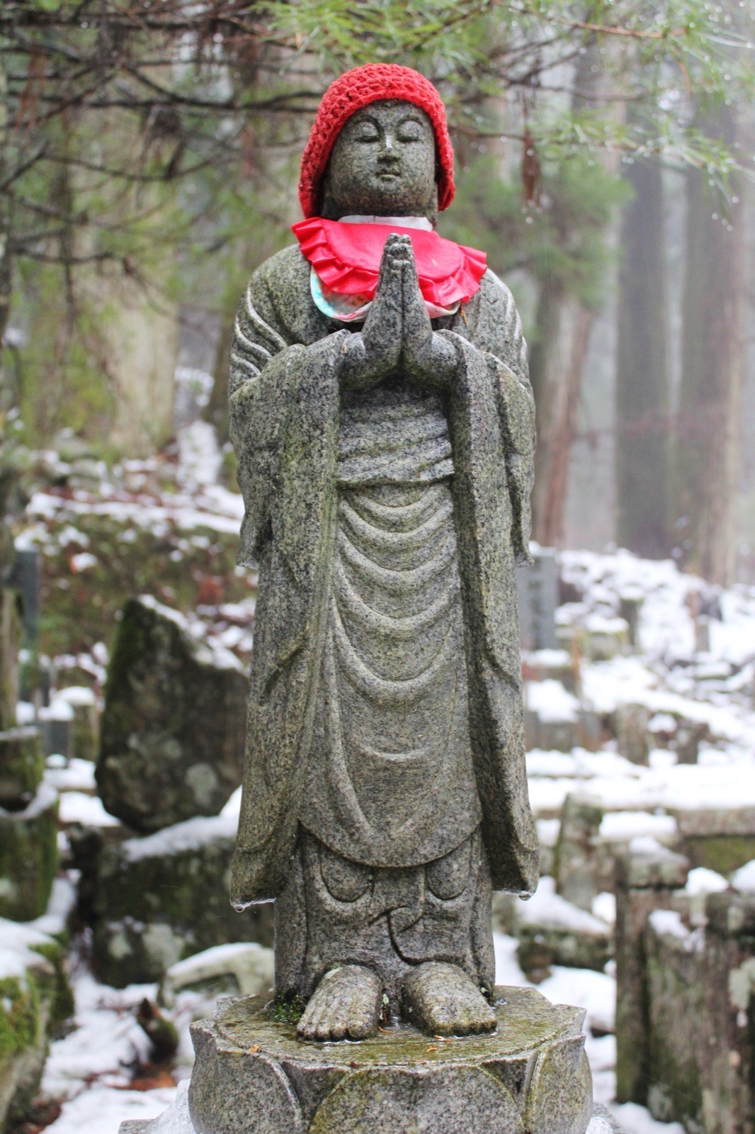 A stone Jizo statue wearing a hand-knitted red cap and bib, hands pressed together in prayer, snow and graves behind