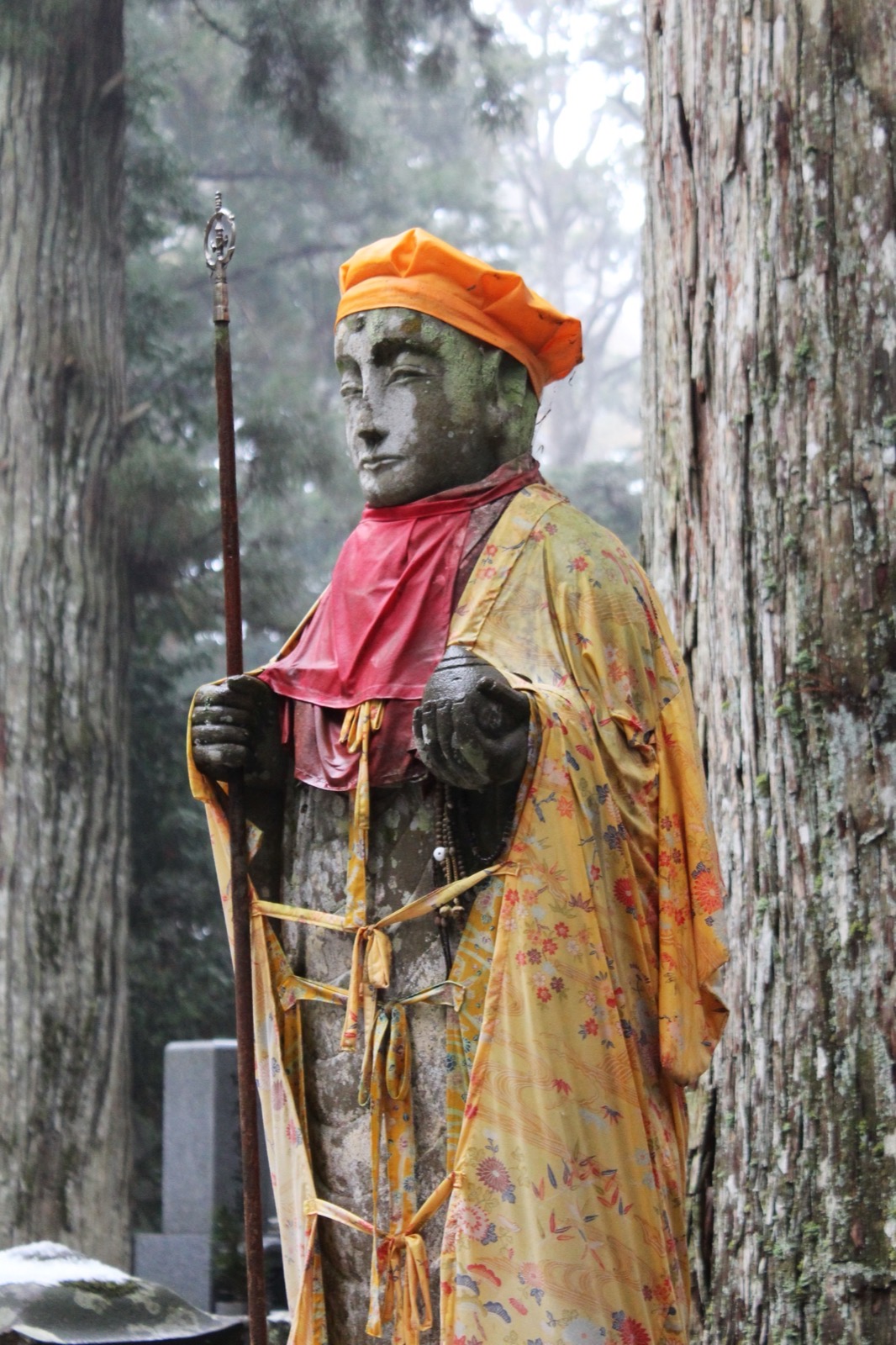 A stone statue dressed in an orange cap, pink bib, and floral kimono, holding a staff among the cedars and graves of Okunoin
