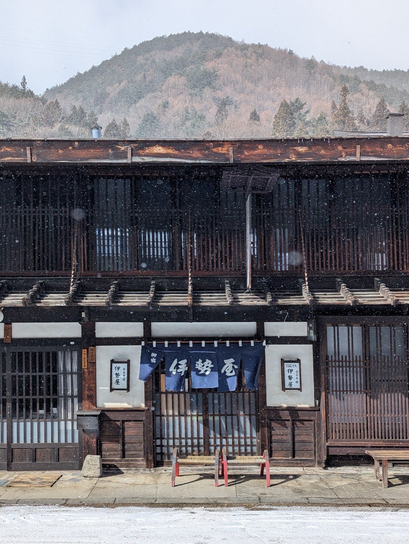 A latticed wooden facade with indigo noren curtains and snow falling against dark timber in Narai-juku