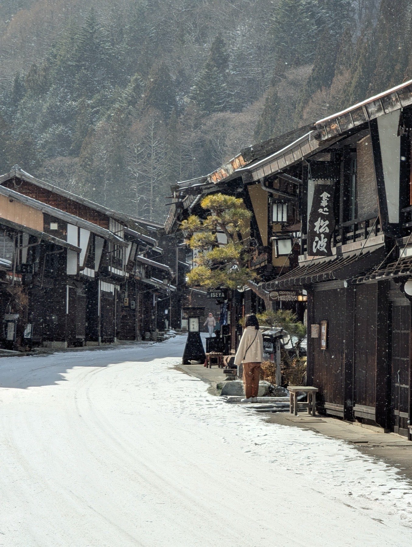 A lone figure walking through heavy snowfall past dark wooden buildings and a pine tree in Narai-juku