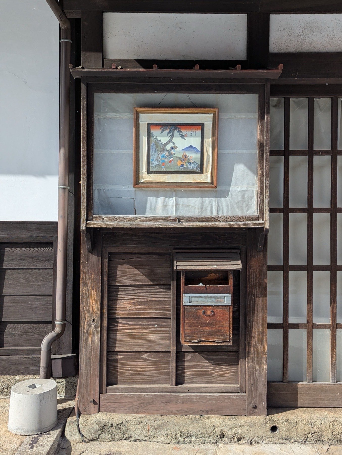 A framed ukiyo-e print displayed in a glass case above a wooden letterbox on the front of a traditional building in Narai-juku