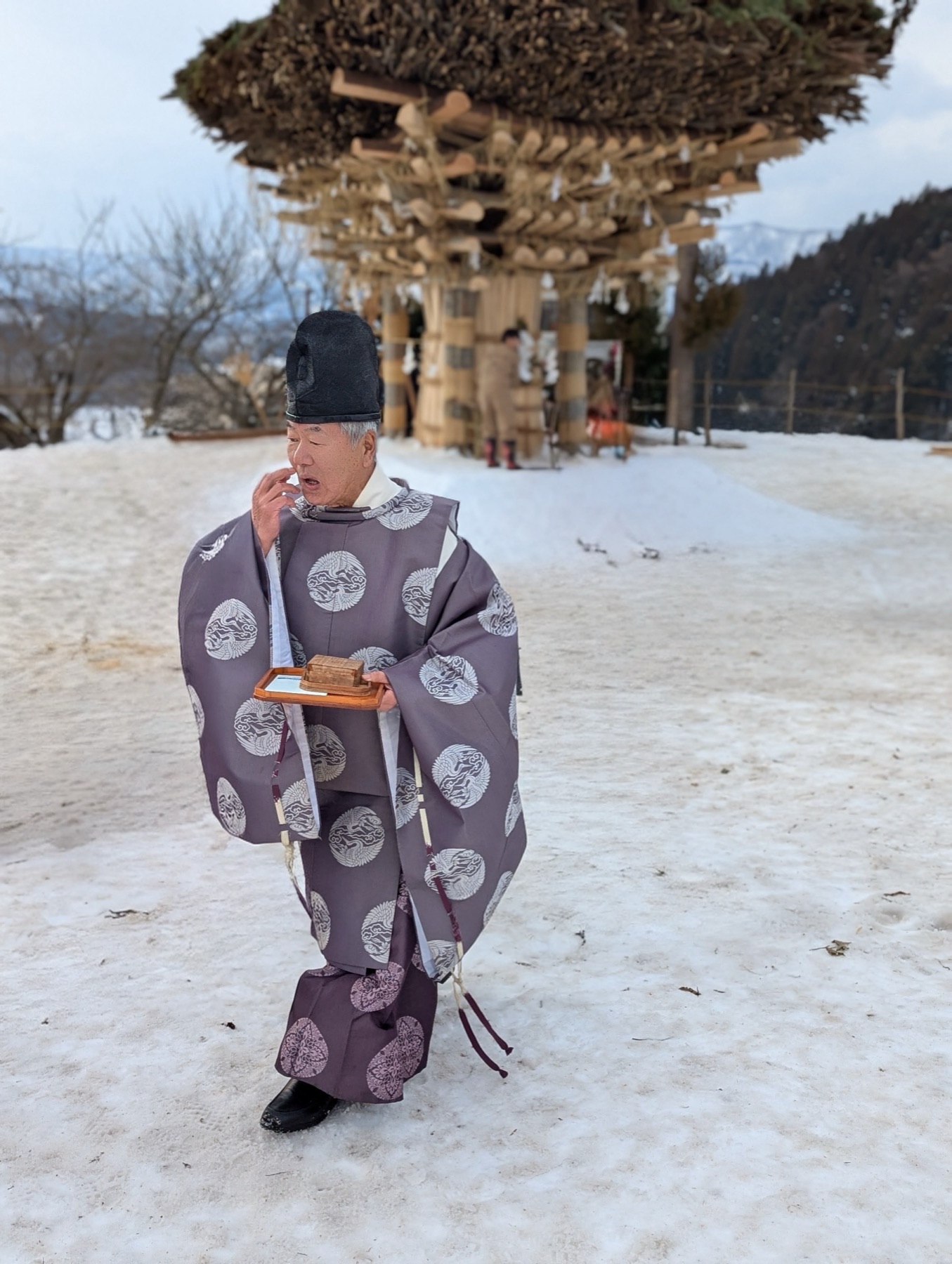 A Shinto priest in grey ceremonial robes walking across the snow at Nozawa Onsen, the wooden shaden shrine rising behind him