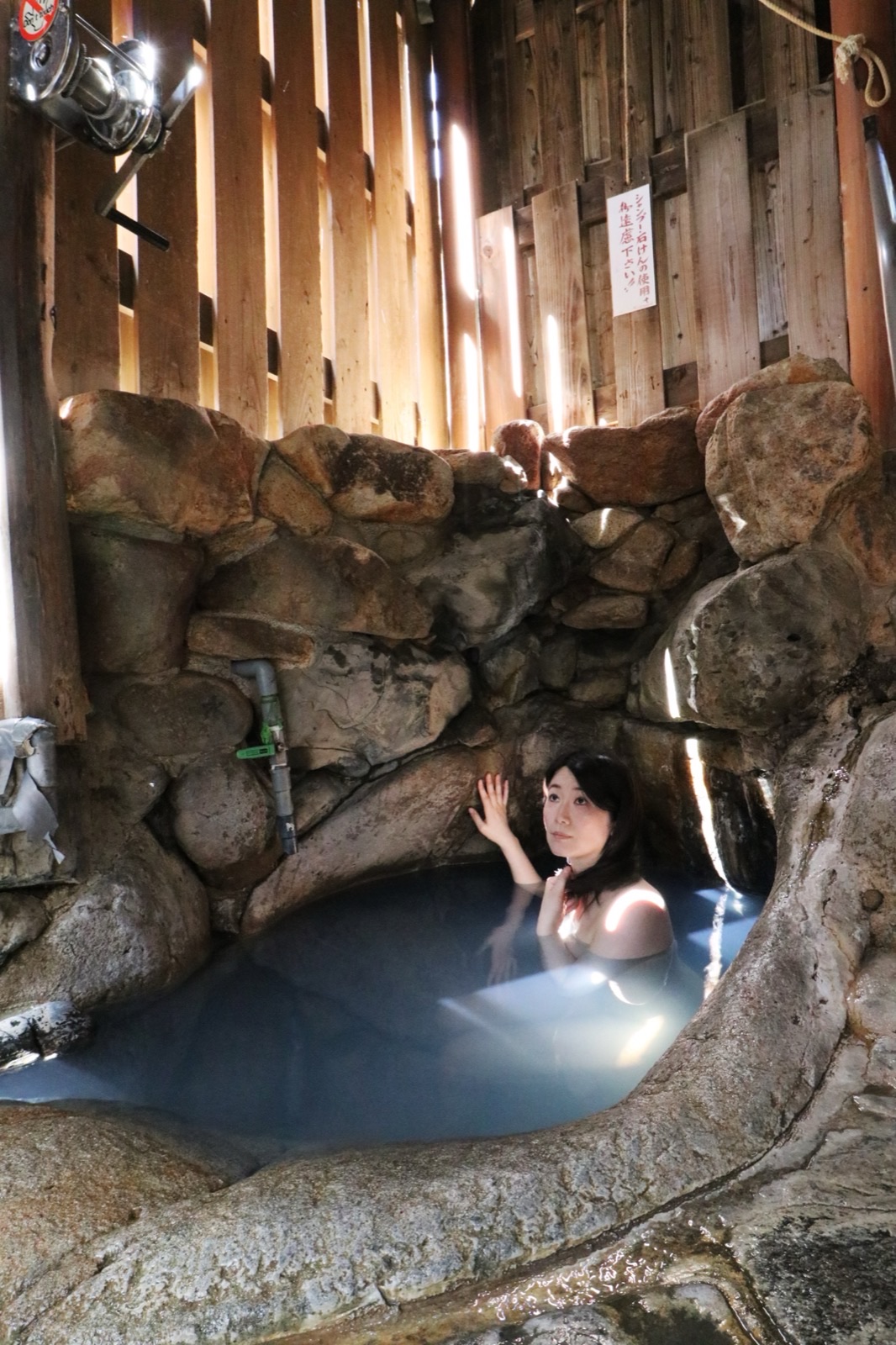 A woman soaking in a natural rock onsen bath, warm light filtering through wooden slats above