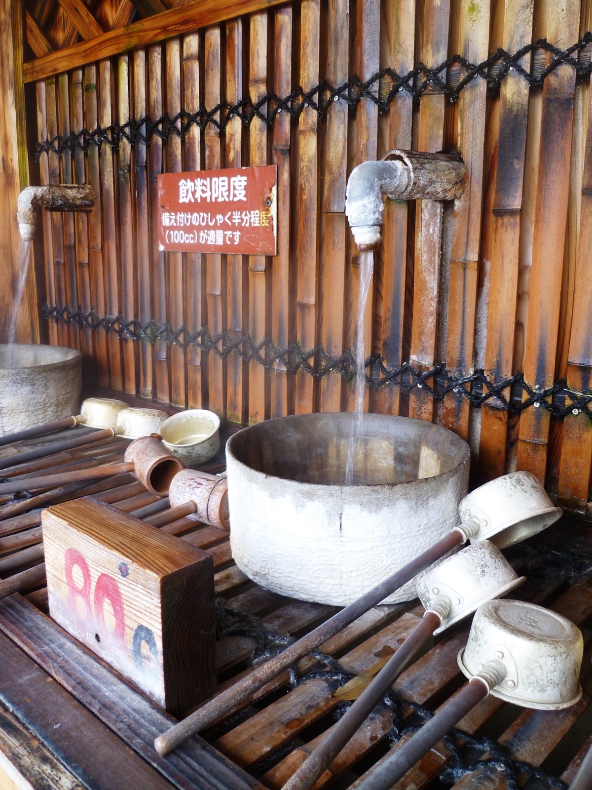 Wooden ladles and a metal basin at an onsen drinking water station, steam rising, bamboo wall behind
